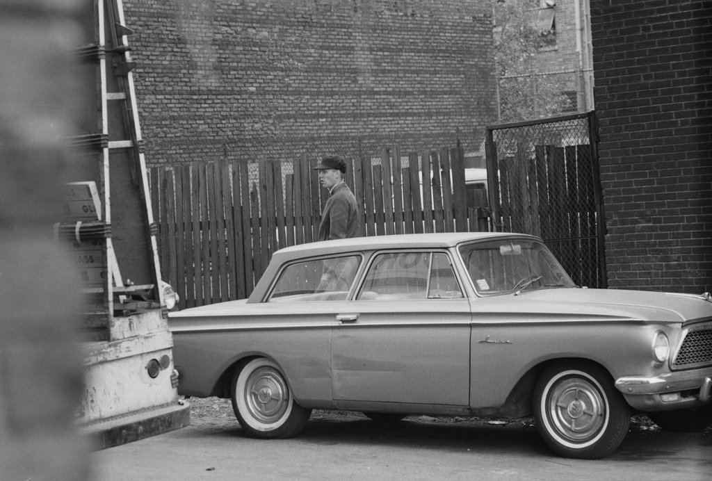 Person standing on the sidewalk between a car and a wooden fence - Haun, Declan, 1937-1994