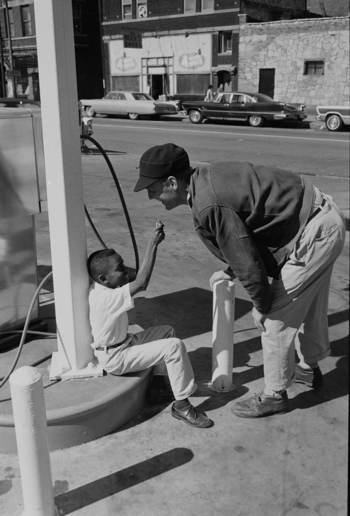 Child playfully aiming gas station air pump hose at a person - Haun, Declan, 1937-1994