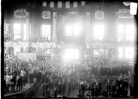 Crowd of people standing on a balcony and on the floor of a large room in the Board of Trade decorated with flags and banners commemorating President Lincoln, there are four men standing in front of a balcony window on the left side of the image - Chicago Daily News, Inc.