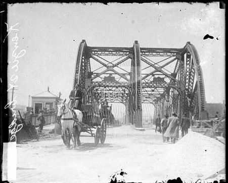 Kinzie Street Bridge with piles of construction material on either side of the approach and men standing in one lane looking at the bridge and a horse drawn wagon being driven away from the bridge in the other lane, viewed from the approach - Chicago Daily News, Inc.
