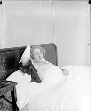 Miss Helen Lambert, lying in a bed in a room, touching the back of her neck with her right hand - Chicago Daily News, Inc.