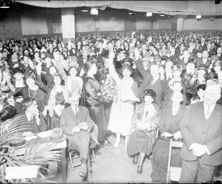 Evangelist Aimee Semple McPherson holding up her right hand, standing with the audience in a seating area in an auditorium - Chicago Daily News, Inc.
