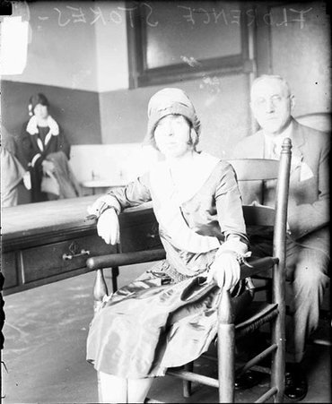 Florence Stokes looking toward the camera, sitting in a chair next to a table, with an unidentified man sitting behind her - Chicago Daily News, Inc.