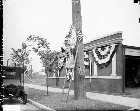 William H. Kleppinger looking toward the camera while standing on a ladder and nailing American flags and decorations on to a telephone pole in East Chicago, Indiana - Chicago Daily News, Inc.