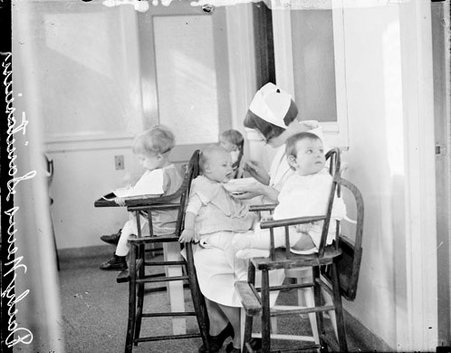 Nurse feeding a baby on her lap while three other babies are sitting in high chairs around her at the Chicago Daily News Sanitarium - Chicago Daily News, Inc.