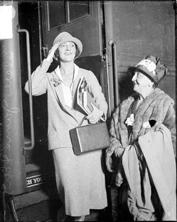 Elsie Janis saluting, standing on the steps of a train car next to her mother who is standing on the ground - Chicago Daily News, Inc.