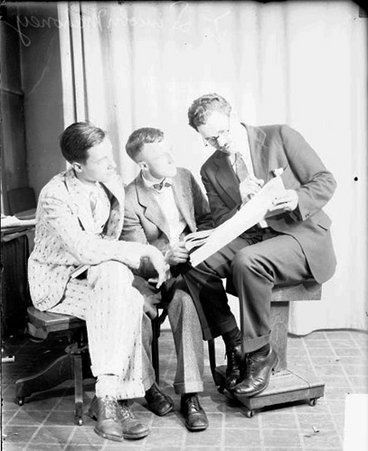 Lincoln Mahoney holding a large paper while sitting with two men in front of a light-colored backdrop - Chicago Daily News, Inc.