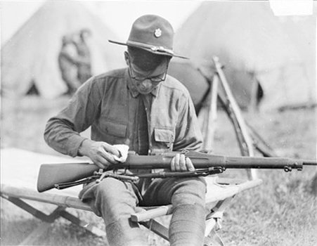Soldier in the Thirty-third Division cleaning a riffle while sitting outdoors on a cot with tents in the background - Chicago Daily News, Inc.