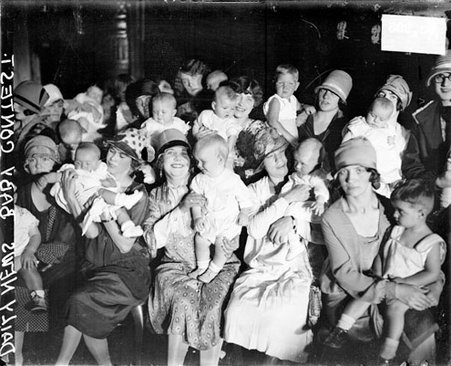 Group of women holding babies, sitting and standing beneath a spotlight in a room, woman in center smiling toward the camera - Chicago Daily News, Inc.