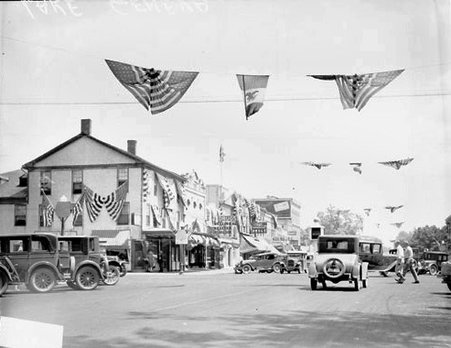 American flags and patriotic banners decorating a commercial street in Lake Geneva, with automobiles driving along the street - Chicago Daily News, Inc.