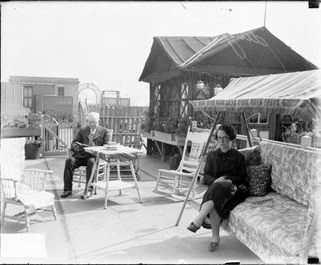 Michael Zimmer reading a book, sitting in a chair on a patio near his daughter who is sitting on a sofa - Chicago Daily News, Inc.