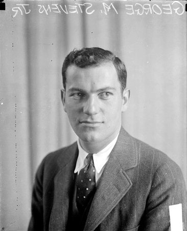 George M. Stevens, Jr. looking to his left, sitting in front of a backdrop in a room - Chicago Daily News, Inc.