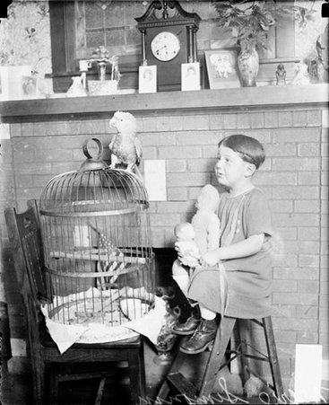 Marie Simonin sitting on a stool next to a parrot standing on top of a bird cage, looking at the parrot - Chicago Daily News, Inc.