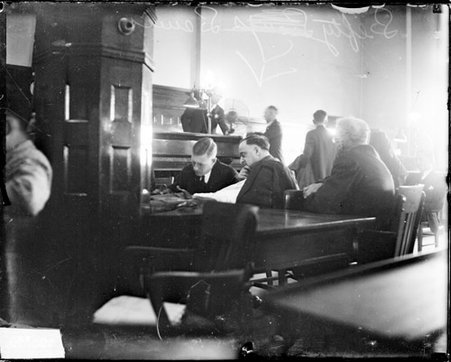 Lefty Lewis sitting in profile at a table in a courtroom, unidentified men are sitting and standing in the room - Chicago Daily News, Inc.