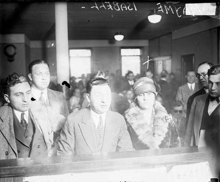 Mayme Isabell standing in front of a wide podium in a crowded courtroom, men surrounding her - Chicago Daily News, Inc.