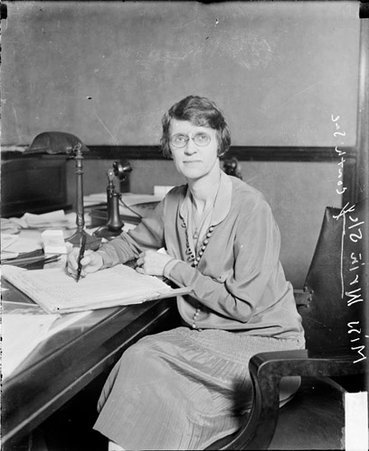 Miss Grinsley looking to her left, holding a pen, sitting at t desk in a room, dark exposure - Chicago Daily News, Inc.
