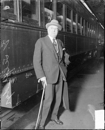 H. G. Selfridge standing with a cane in front of a passenger train car in a train station - Chicago Daily News, Inc.