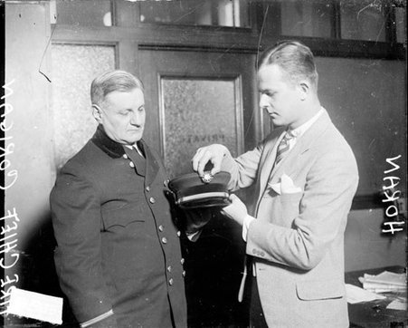 Michael Corrigan and Mr. Horan standing in an office, Corrigan holding his uniform cap, Horan placing a button on it - Chicago Daily News, Inc.