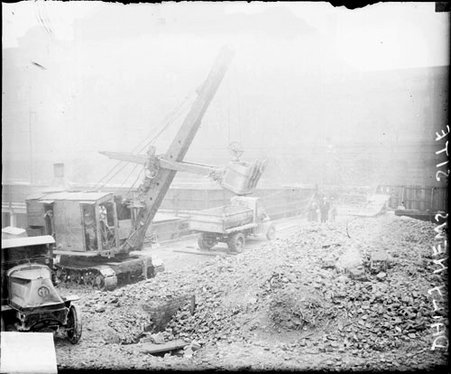 Three people standing on and a truck driving over a bridge, tractor standing in the foreground at a construction site - Chicago Daily News, Inc.