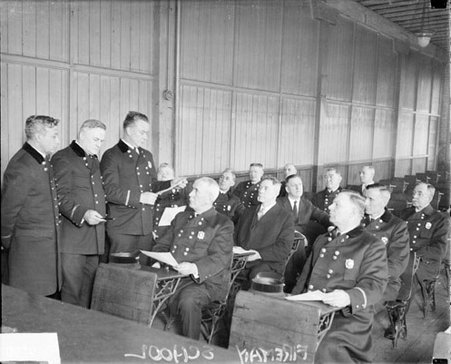 Firemen sitting in desks in a room, three fireman standing at the front of the room, dark exposure - Chicago Daily News, Inc.