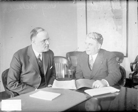 Oscar E. Nelson and Guy Richardson looking at one another, sitting at a table in a room - Chicago Daily News, Inc.