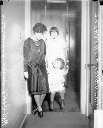 Mrs. James Herbert Mitchell standing in a doorway in a house with two young girls, one sitting in a swing, dark exposure - Chicago Daily News, Inc.
