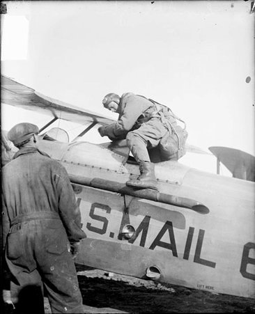 Charles A. Lindbergh climbing into the cockpit of a propeller airplane on a field, mechanic standing in front of the plane - Chicago Daily News, Inc.