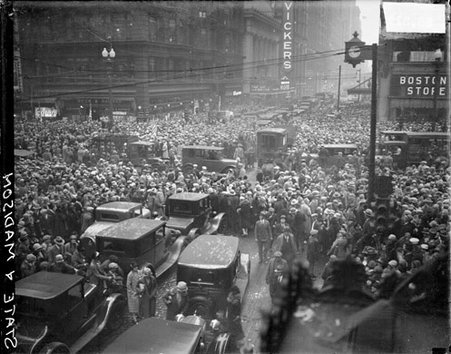 Automobiles passing through large crowds standing on State and Madison Streets, looking west on Madison from State - Chicago Daily News, Inc.