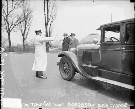 Police officer George Ferguson standing in front of an automobile on a street, two pedestrians in background - Chicago Daily News, Inc.