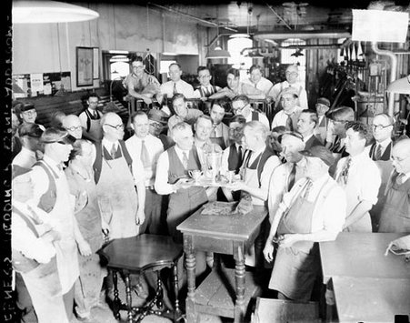 Two men holding a tray bearing chalices, standing amid a crowd of workers in a factory room - Chicago Daily News, Inc.