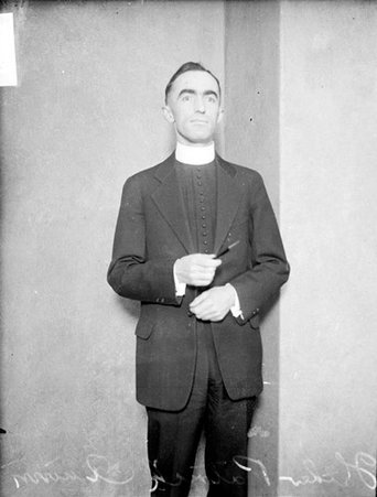 Father Patrick Quinn looking toward the camera, standing in front of a backdrop in a room - Chicago Daily News, Inc.