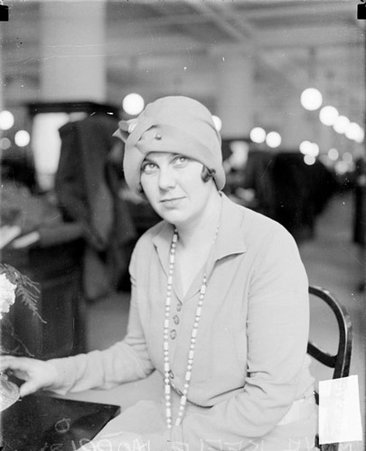 Anna K. Morris looking up and to her left, sitting at a table in a large room - Chicago Daily News, Inc.