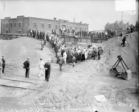 Crowd standing on and below small sand hills, construction equipment in foreground - Chicago Daily News, Inc.