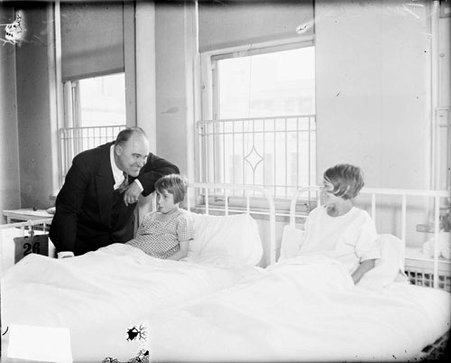 Man smiling at and standing next to two young girls sitting up in beds in a room - Chicago Daily News, Inc.