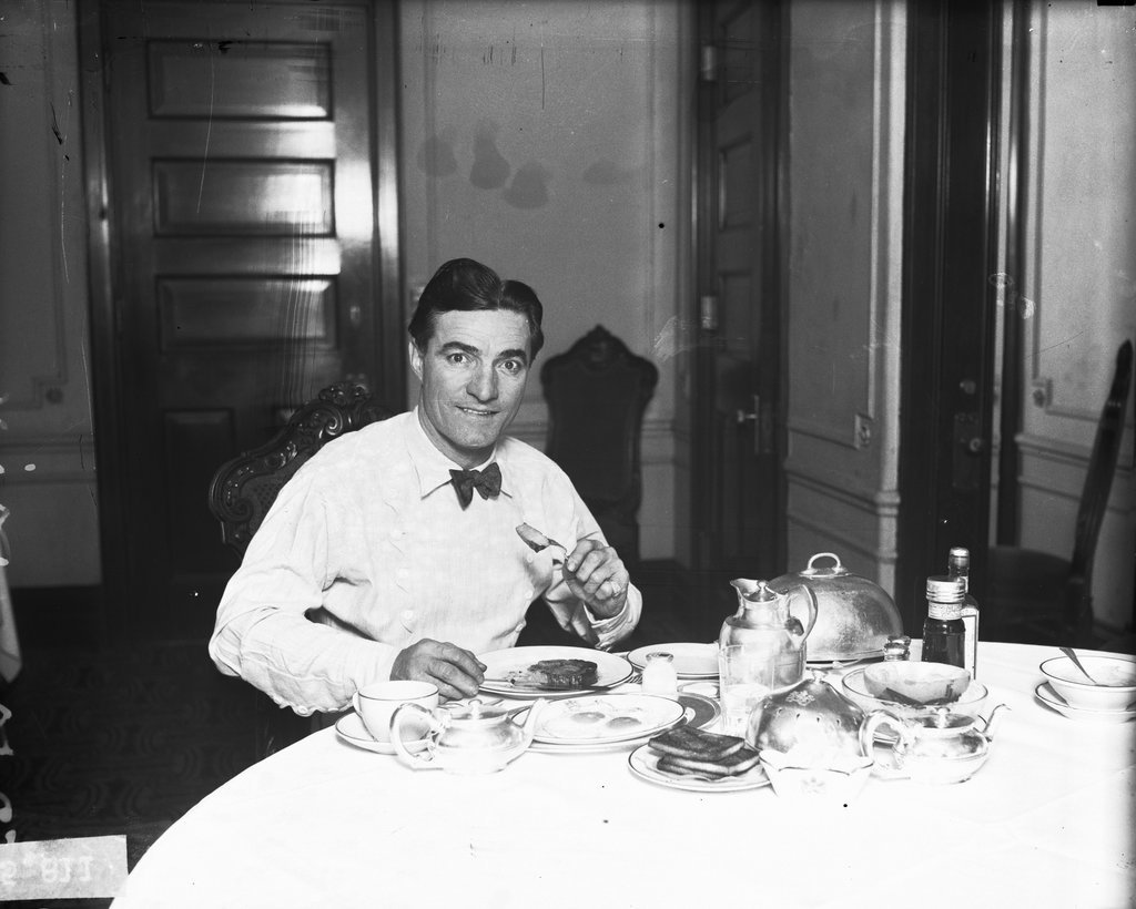 Actor Tom Mix sitting at a table set for breakfast in a room - Chicago Daily News, Inc.