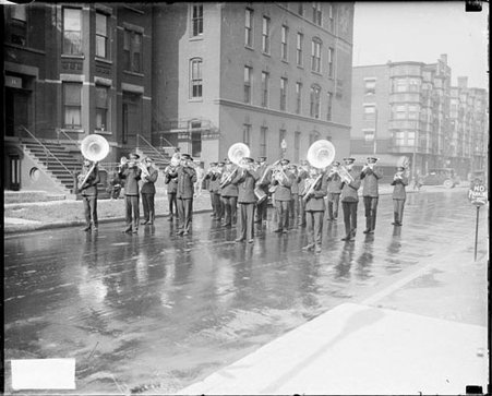 Salvation Army marching band playing instruments, standing on a wet street in front of a building - Chicago Daily News, Inc.