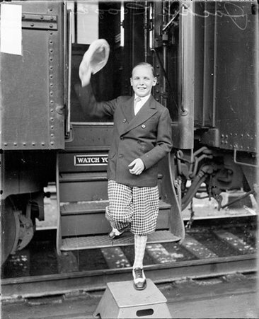 Jackie Coogan holding up his hat, standing in front of a passenger train car in a railroad station - Chicago Daily News, Inc.