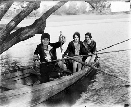 Young woman holding a fish on a fishing line, sitting in a rowboat in a body of water with two other women - Chicago Daily News, Inc.