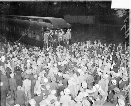 Alfred E. Smith standing with a group of people on the rear platform of a passenger train car in a railroad station - Chicago Daily News, Inc.