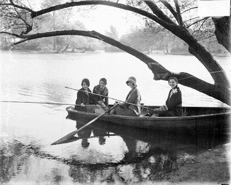 Four young women sitting in a rowboat in a body of water, two women holding fishing poles - Chicago Daily News, Inc.