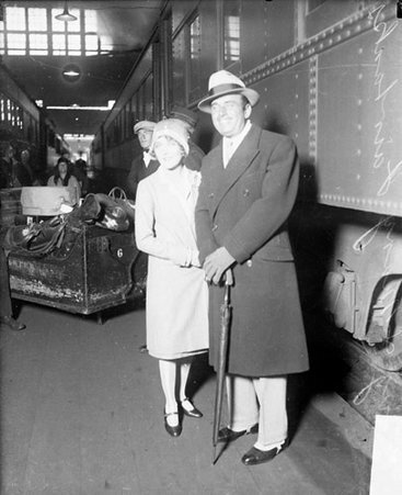 Actors Douglas Fairbanks and Mary Pickford standing in front of a passenger train car in a railroad station, full luggage cart is visible in the background - Chicago Daily News, Inc.