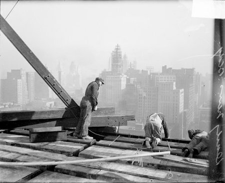 Man standing high atop the steel frame of the new Chicago Daily News building looking down at two men who are leaning over the edge of the frame - Chicago Daily News, Inc.