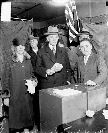 Charles G. Dawes, Vice-President of the United States from 1925-1929, and his wife, nee Caro D. Blymyer, holding ballots, standing behind a ballot box at a voting poll center in a room, dark exposure - Chicago Daily News, Inc.