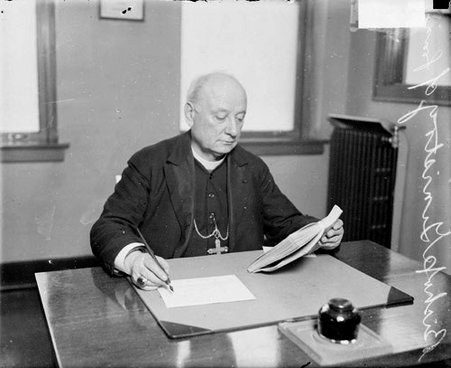 Bishop Ginistry of France sitting at a desk in a room in Chicago, Illinois, holding a book and writing on a small piece of paper - Chicago Daily News, Inc.