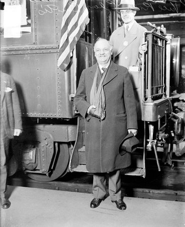 Charles Curtis, U. S. looking to his right, standing in front of a passenger train car in a railroad station, an American flag is partially visible in the background - Chicago Daily News, Inc.