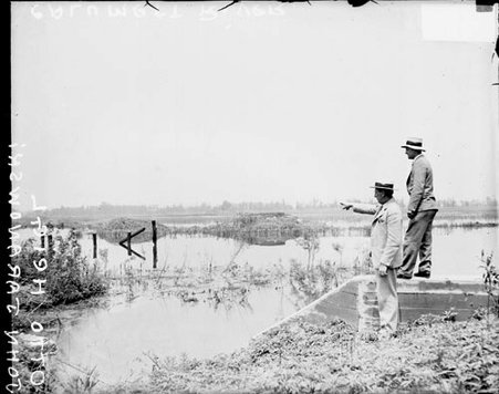 Calumet Mayor John Jaranowski and Otto Heigl standing on shore of flooded Calumet River at Burnham, Illinois - Chicago Daily News, Inc.