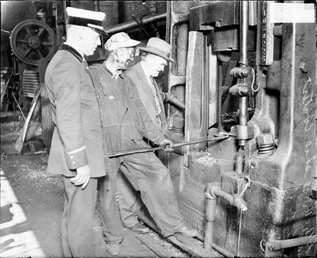 Man covered in grease and wearing work clothes holding a metal pole to a machine - Chicago Daily News, Inc.