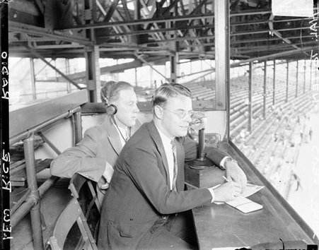Hal Totten sportswriter and announcer for WMAQ radio station with Lew Rice sitting in the announcer's stand and a microphone is sitting on the counter in front of him in an outdoor stadium - Chicago Daily News, Inc.