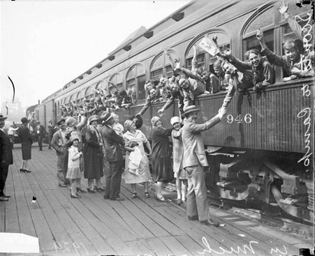 Boy Scouts leaning and waving at a group of adults out of the windows of a passenger train car in a railroad station leaving on a trip to Michigan, dark exposure - Chicago Daily News, Inc.