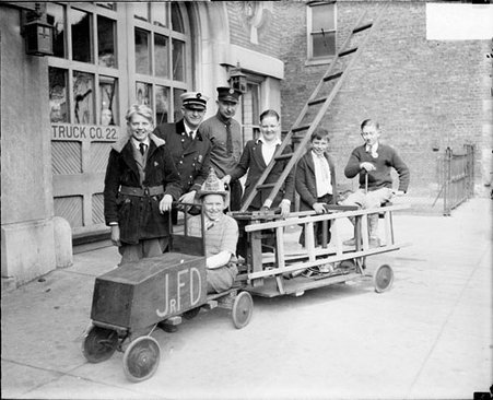 Boy wearing a firefighter's helmet, sitting in a miniature wooden fire truck in front of a fire station - Chicago Daily News, Inc.